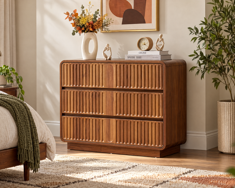 A mid-century modern wooden chest of drawers in a warm medium brown finish, styled in a bright bedroom next to a bed and a potted plant.