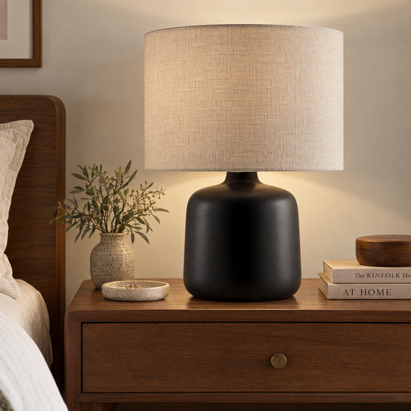 A modern table lamp with a matte black ceramic base and a textured beige linen drum shade, illuminated on a wooden nightstand in a cozy bedroom setting.