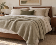 A styled bedroom scene showing a bed dressed in white bedding with a textured light beige throw blanket draped across the foot, a dark wood bed frame, matching pillows, bedside tables, a lamp, and a window with natural light.