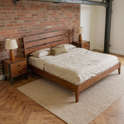 Acacia Wood Slat Bed Frame with ivory bedding and a camel throw, shown at an angle in a warm brick and steel loft bedroom with a flat oatmeal rug