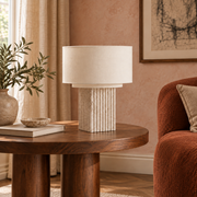 Fluted square travertine table lamp with a cream drum shade on a walnut table in an Artisan living room with textured plaster and rust upholstery