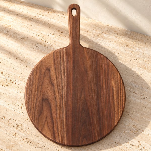 Walnut Round Paddle Cutting Board — Small 12" Circle on a sunlit pale travertine counter