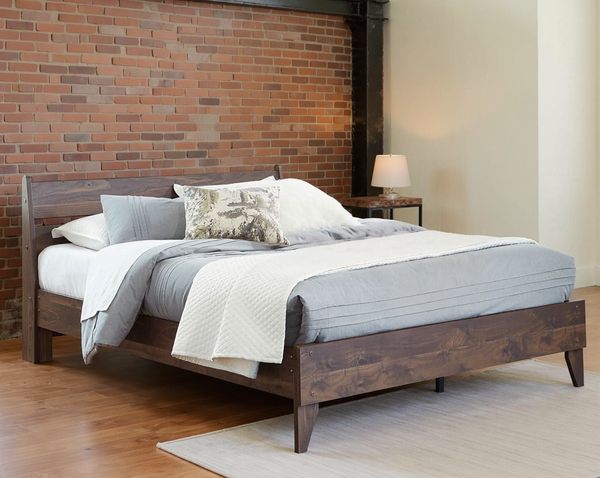 Dark brown wood-grain bed with a slatted headboard and layered pale bedding in a brick loft bedroom.