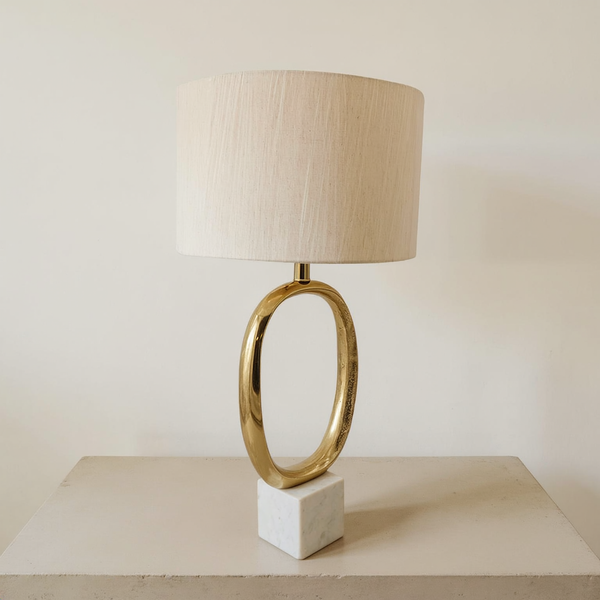 Oval gold metal table lamp with a linen drum shade and marble block base on a neutral tabletop
