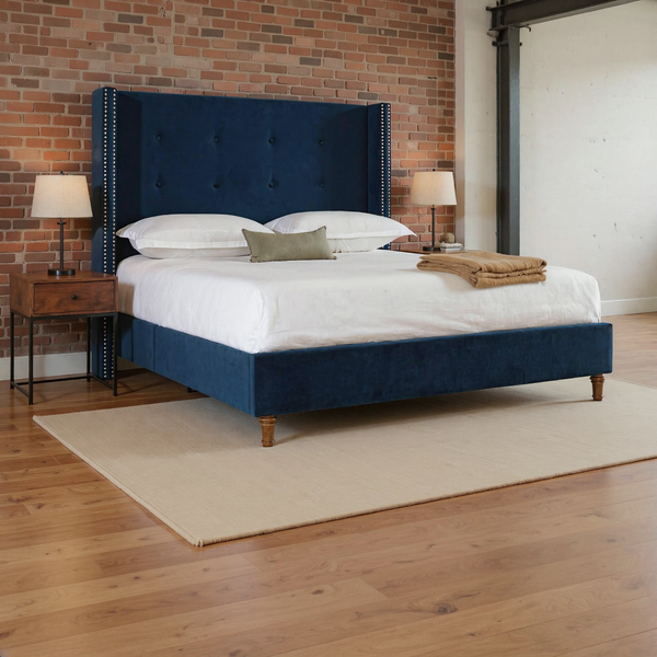 Navy tufted velvet wingback queen bed with nailhead trim and turned wooden feet, ivory bedding and camel throw in a warm brick loft bedroom on a flat oatmeal rug.