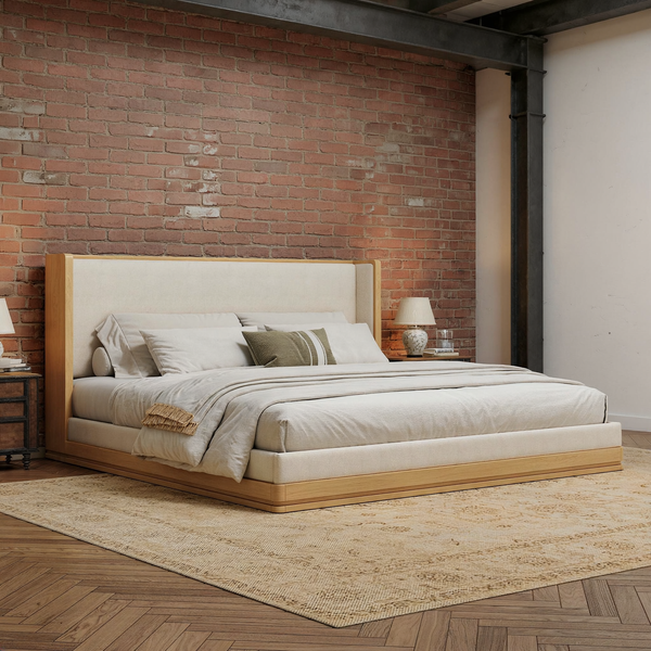 Light wood and cream upholstered platform bed with ivory bedding in a brick and steel loft bedroom, shown at an angle with a flat oatmeal rug.